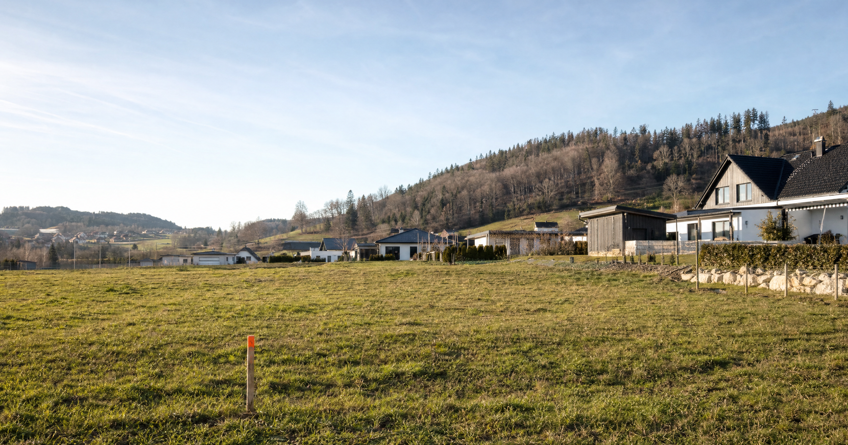 Unbebautes Baugrundstück mit Vermessungspflock und Wohnhäusern im Hintergrund